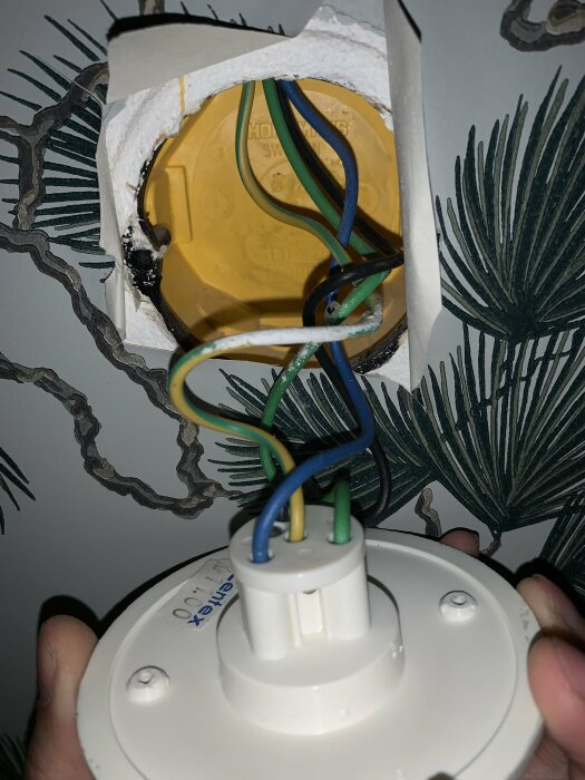 Installation av ljusarmatur, öppen kabelanslutning, vägguttag, ornamenterad tapet, elektriskt arbete pågår.