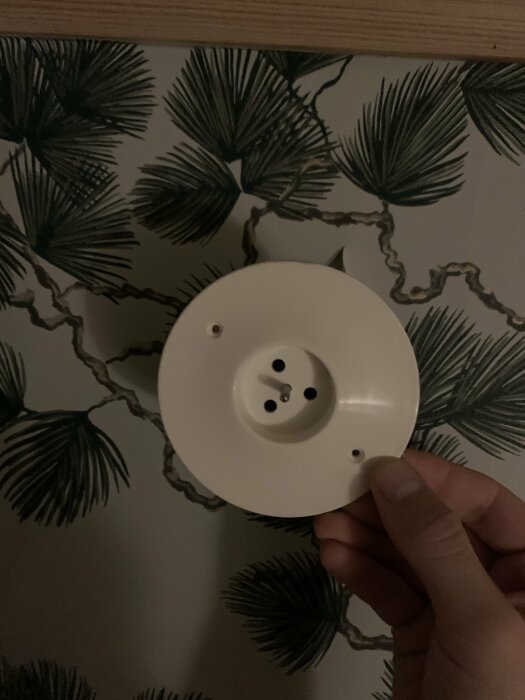 En hand som håller en vit rund rökdetektor framför en tapet med tropiskt palmbladsmönster.