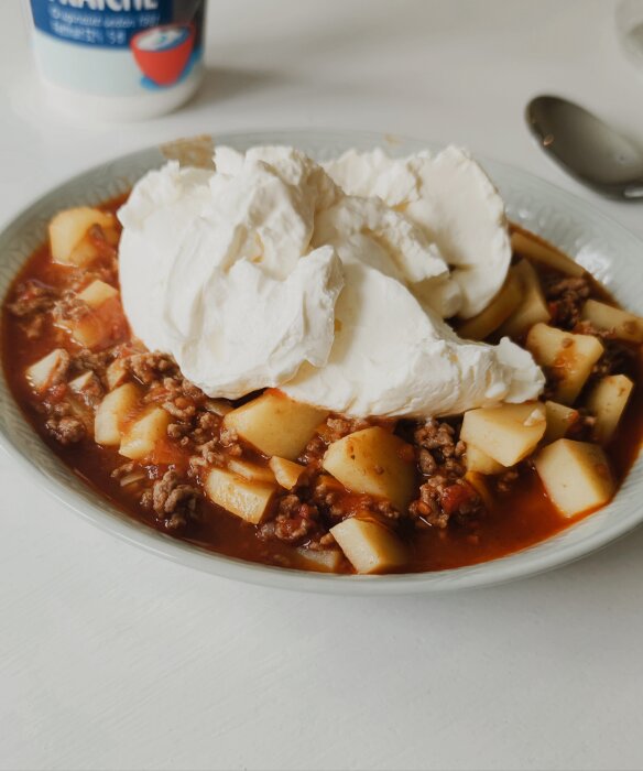 En tallrik chili med köttfärs, potatis och en stor klick gräddfil, med en sked och behållare i bakgrunden.