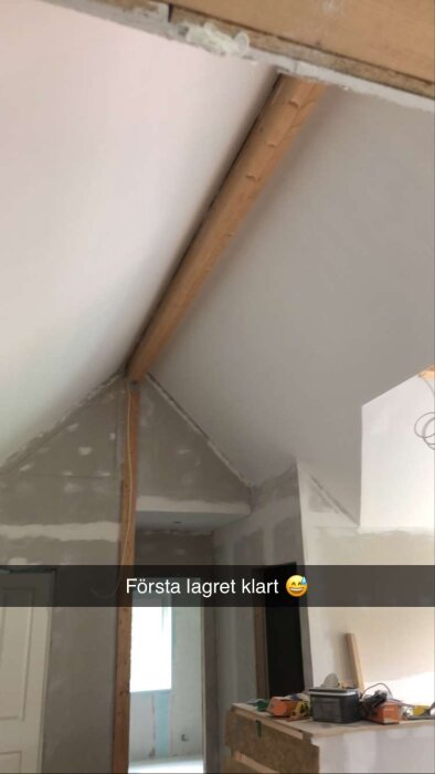 Renoveringsarbete, taket och väggen delvis spacklade, träbjälkar synliga, byggmaterial och verktyg, text "Första lagret klart".