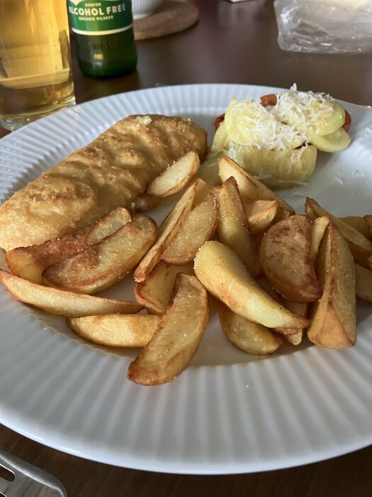 Friterad fisk, klyftpotatis, potatis med riven ost, alkoholfri öl i bakgrunden; måltid på vit tallrik.