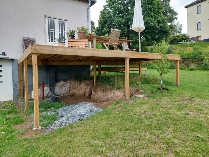 Trädäck under konstruktion vid ett hus, stödstolpar, trädgårdsmöbler ovanpå, utsikt mot trädgård och andra hus.