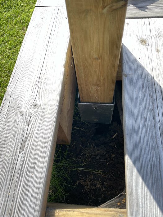 Trästolpe i metallhållare på gräs, mellan gråa träbrädor, solljus, skugga, jord nertill.