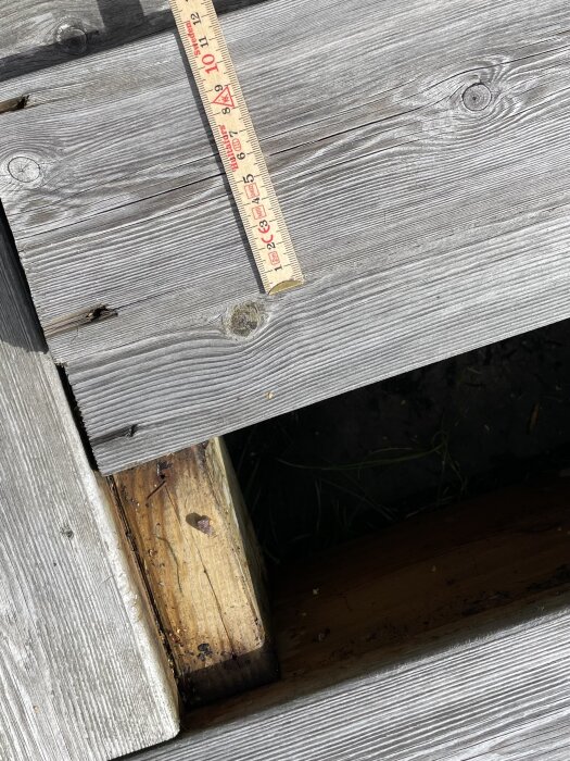 Trämätsticka på grå träyta bredvid öppning med synlig brun träbjälke och mörk utrymme under.