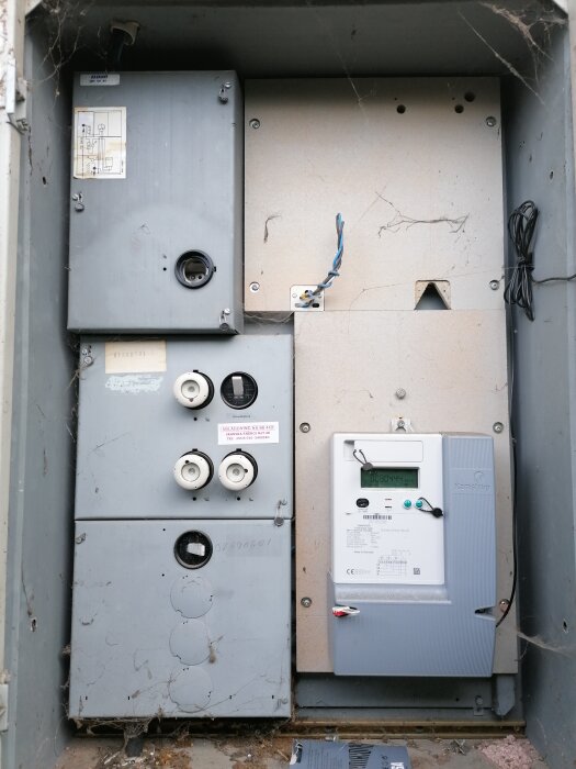 Gammal och ny elräkneverk i en dammig, spindelvävsfylld utomhuselektricitetsbox, slitna kablar, slitna etiketter.
