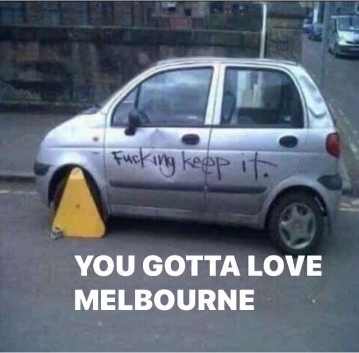 En parkerad bil med klotter och en trafikkotte som hindrar bogsering, text skämtar om Melbourne.