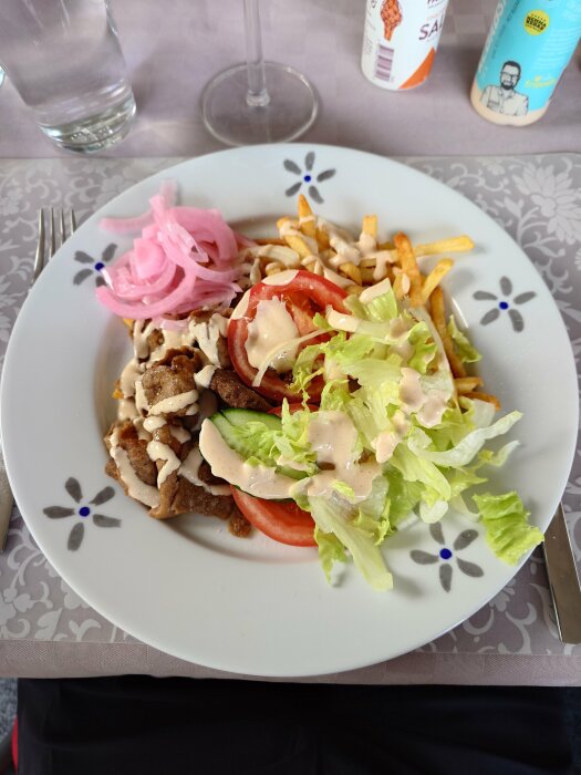 En tallrik med kebabkött, pommes frites, sallad, tomat, inlagd rödlök och dressing. Dryck i bakgrunden.