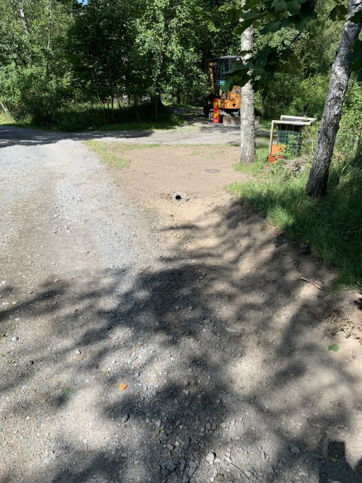 Grusväg, träd, skuggor, soligt väder, en orangefärgad skogsmaskin i bakgrunden, sommardag.