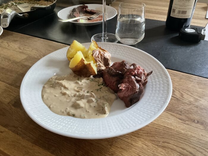 Tallrik med stekt kött, kokt potatis, pepparsås. Bord med vatten, vin, kryddor i bakgrunden.