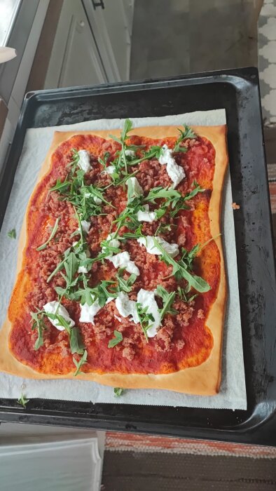 Hemlagad rektangulär pizza med tomatsås, malet kött, ruccola och klumpar av färskost på bakplåt.