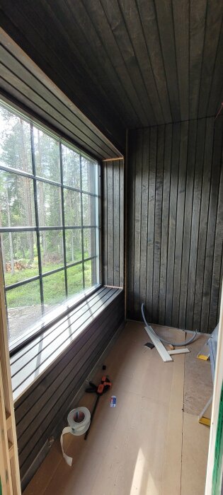 Träbeklädd inomhusrum under konstruktion eller renovering med stora fönster som vetter mot träd.