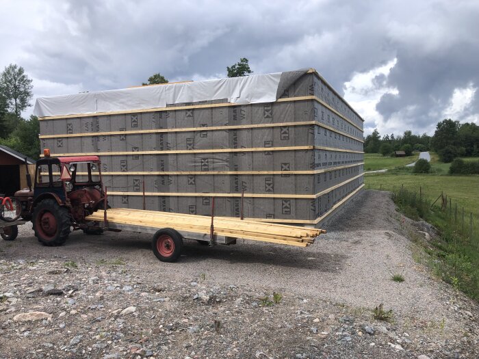 Lastad släpvagn med byggmaterial vid traktor, molnig himmel, grusväg, grönt landskap.