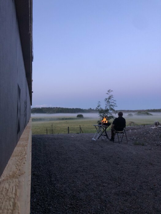 Person sitter framför brasa i dimmig äng vid skymning, lugn atmosfär, utomhus, nära byggnad.