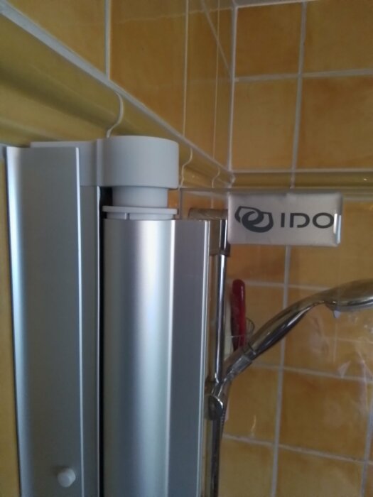 Duschhörna med gula kakelväggar, silverfärgad duschdörr och IDO-logotyp.