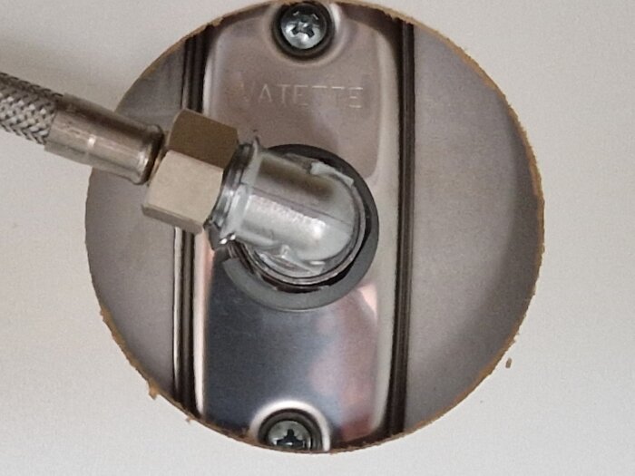 Närbild på en genomföring för en flexibel vattenledning i ett hål. Metalldetaljer, fastsättningselement och vägg syns.