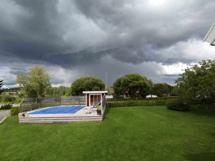 Mörka moln, trädgård med pool, dramatisk himmel, stormväder närmar sig, lugn innan ovädret.