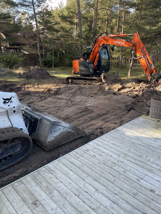 Grävmaskin, skidsteer, grushög, trädäck, skogsmiljö, grävarbete, jordhög, hus i bakgrunden, soligt, gräs.