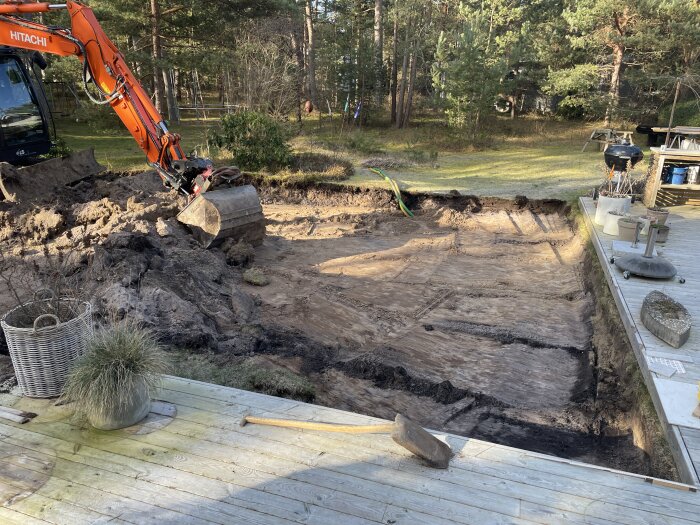 Grävmaskin gräver i trädgård, skapar hål, terrass i förgrunden, skogsmiljö, utomhusbyggnadsarbete under solig himmel.