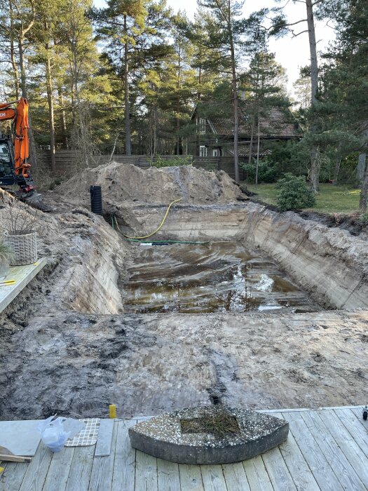 Stor utgrävning nära skog med grävmaskin, vattenansamling, trädäck och villakvarter i bakgrunden. Byggarbetsplats.