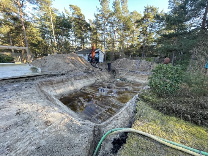 En grävmaskin vid en tom pool under konstruktion eller renovering, omgivet av natur och byggnationer.