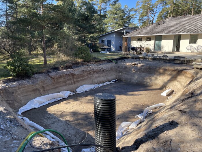 Grävd grop för pool framför hus, trädgård under konstruktion, dukar längs kanten, slang, soligt väder.