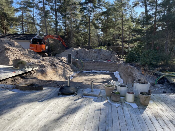 Grävmaskin vid grävarbetsplats, träterrass, planteringskrukor, skogsmiljö, jordhögar, sandsäckar, soligt.