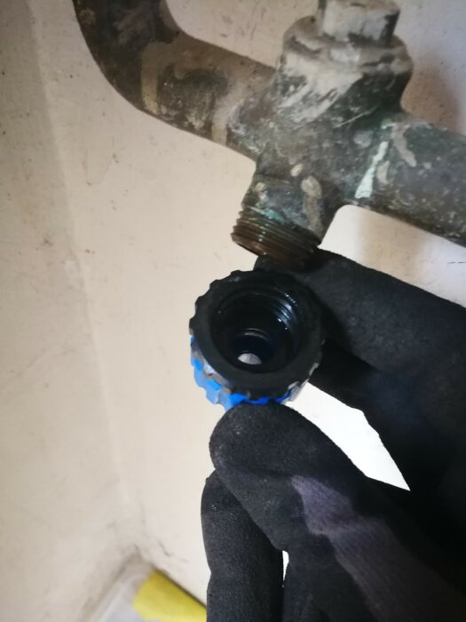 Hand i svart handske håller en blå svart plastkork nära en korroderad vattenkran mot vit vägg.