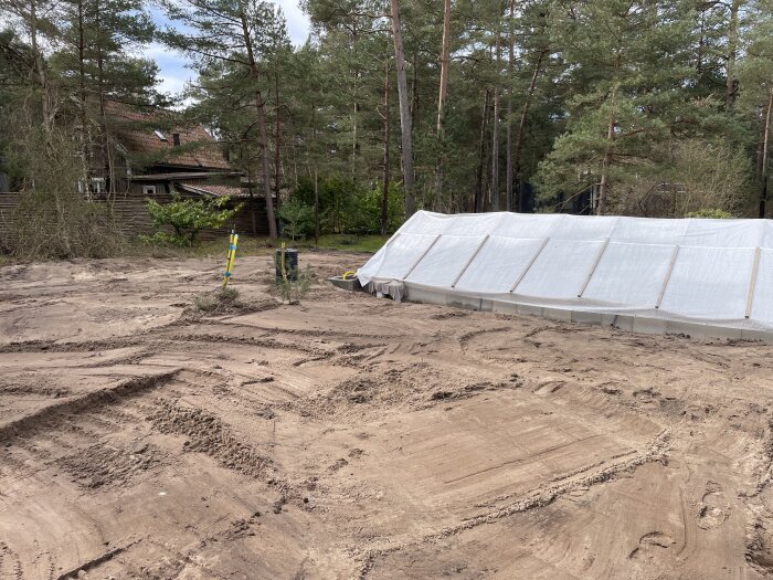 Byggarbetsplats med sand, hus i bakgrunden, vit presenning, träd, molnig himmel.