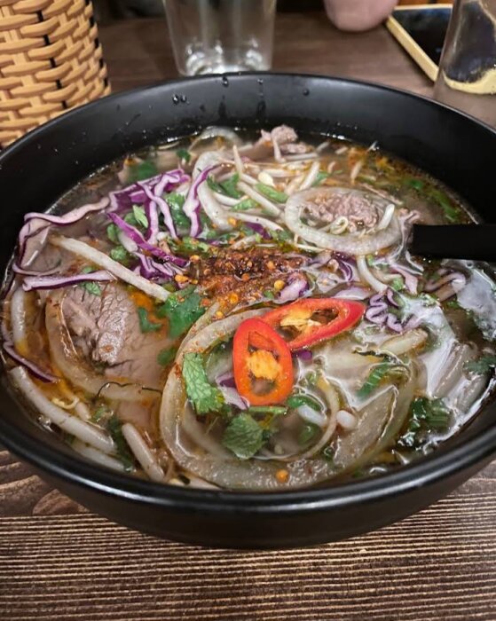 En skål med asiatisk nudelsoppa, troligen pho, med kött, örter och rödlök.