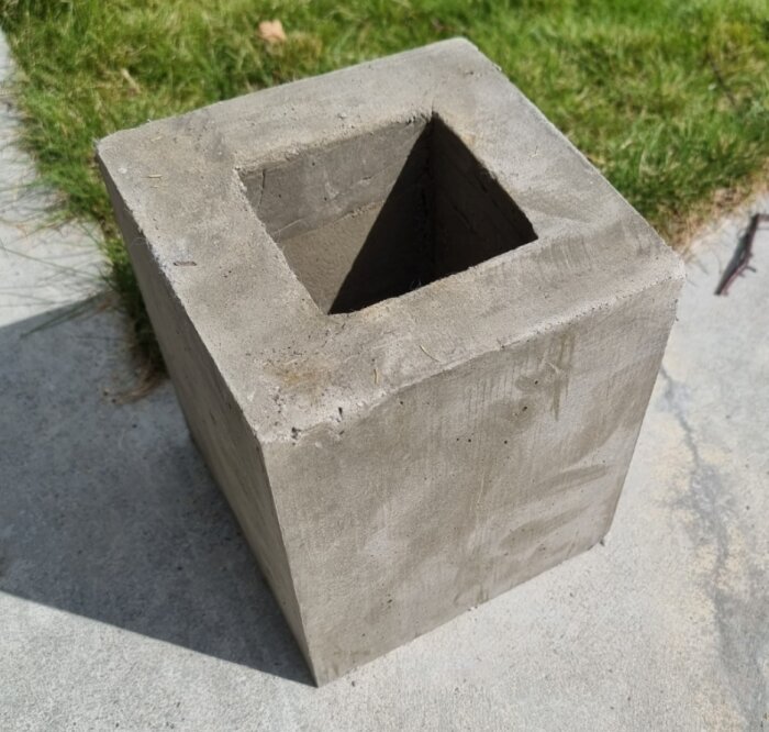 Grå betongblock med ett fyrkantigt hål, placerat på trottoar vid gräs.