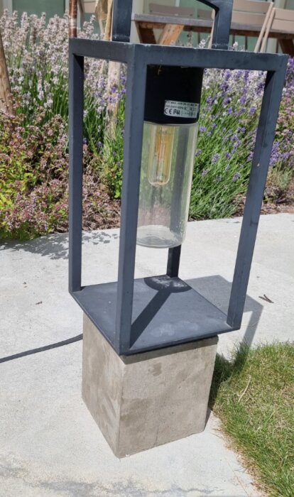 Modern utomhuslykta på betongpelare, genomskinlig ljuskälla, lila blommor i bakgrunden, dagtid.