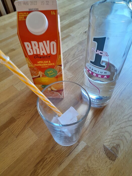 Juicekartong, vodkaglas och sugrör, istärningar i ett dricksglas på träbord.