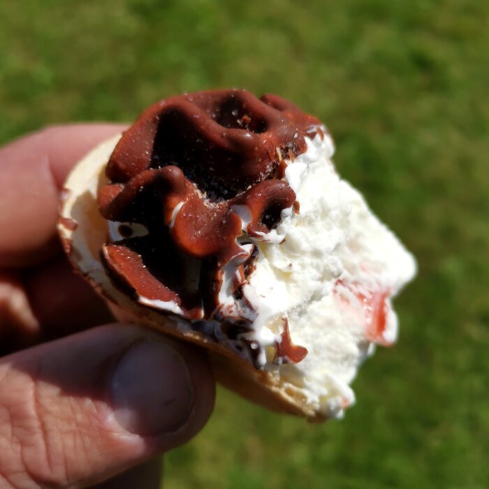 En hand håller en halväten glass med chokladtopping och grädde mot en gräsmatta i bakgrunden.