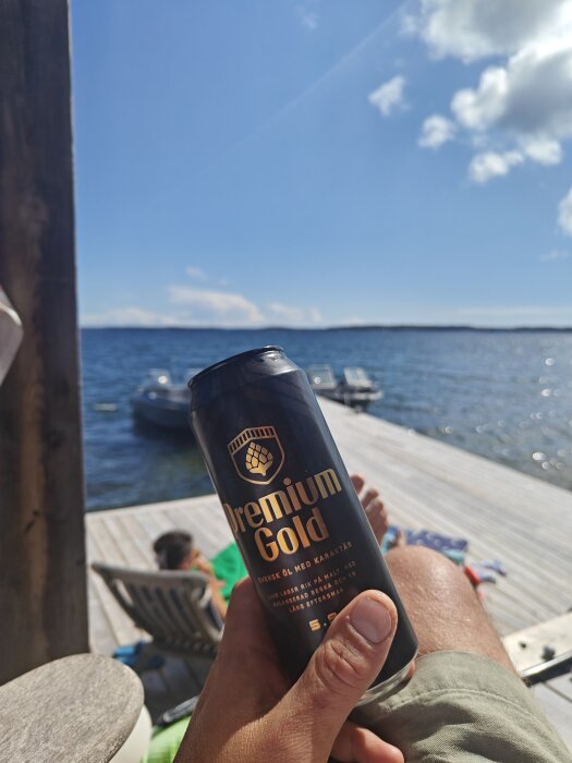 Person håller en ölburk på brygga vid sjö, soligt väder, avkoppling, båtar i bakgrunden.