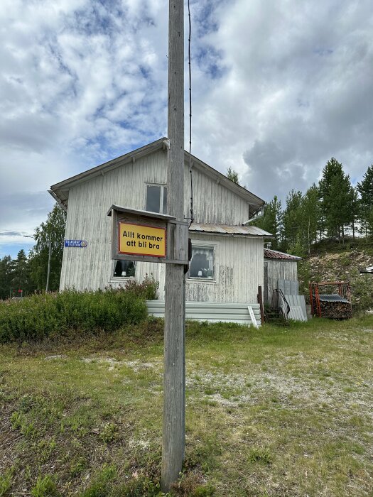 Trähus, telestolpe, skylt med texten "Allt kommer att bli bra", gräs, molnig himmel.