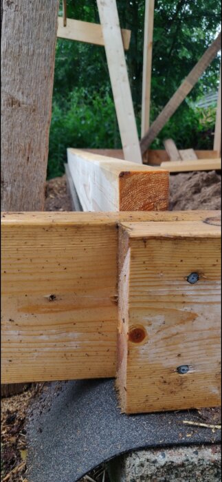 Detaljbild av träkonstruktion; skruvar, kapade träbjälkar och grunden av ett byggprojekt i bakgrunden.
