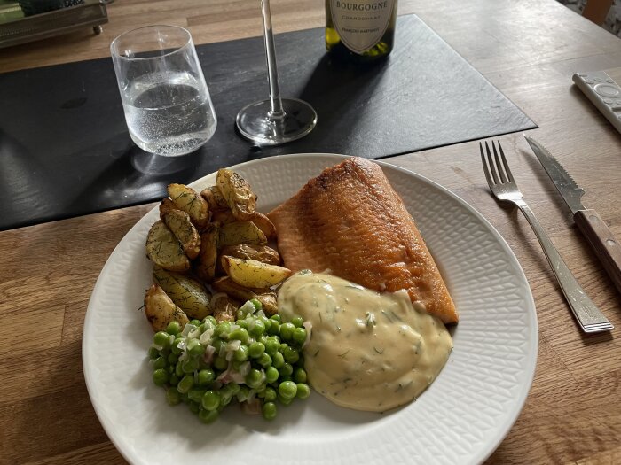 Middagstillfälle med lax, rostad potatis, ärter, sås, vatten, vin, bestick, på ett matbord.