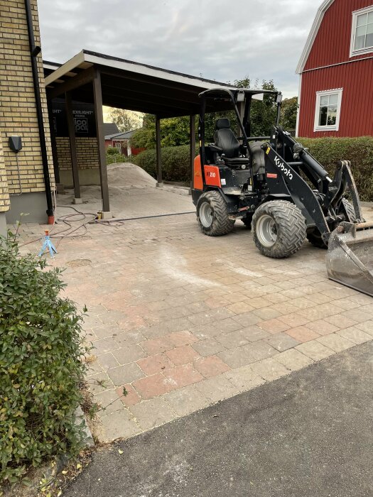 Kompaktlastare framför en tegelbelagd uppfart, carport, hus, byggarbetsplats, hög med material, grå himmel.