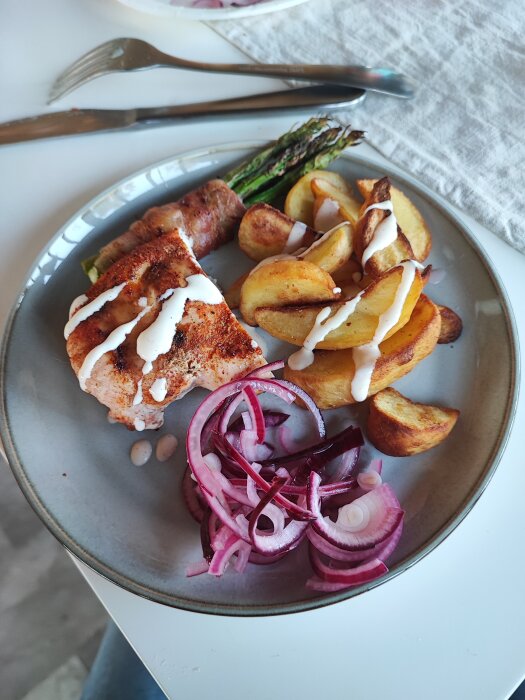 Måltid med grillad kyckling, potatis, sparris och rödlök, serverat på tallrik med bestick.