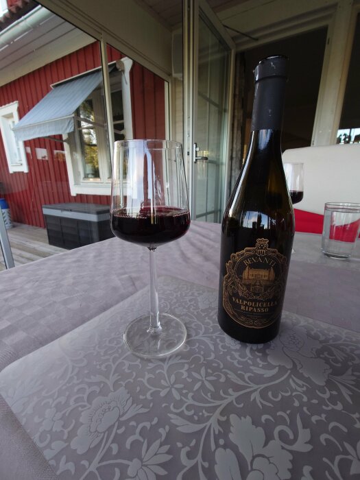 Ett glas rödvin och en vinflaska på ett dukat bord utomhus vid en veranda.