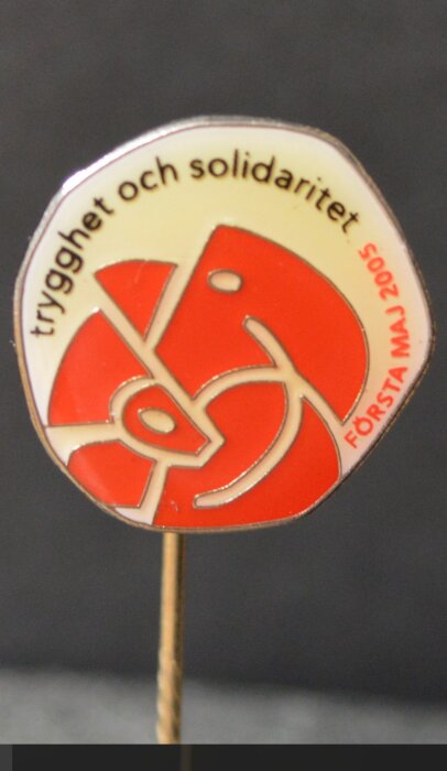 Röd och vit knappnål med text "trygghet och solidaritet. FÖRSTA MAJ 2005" och grafisk design.