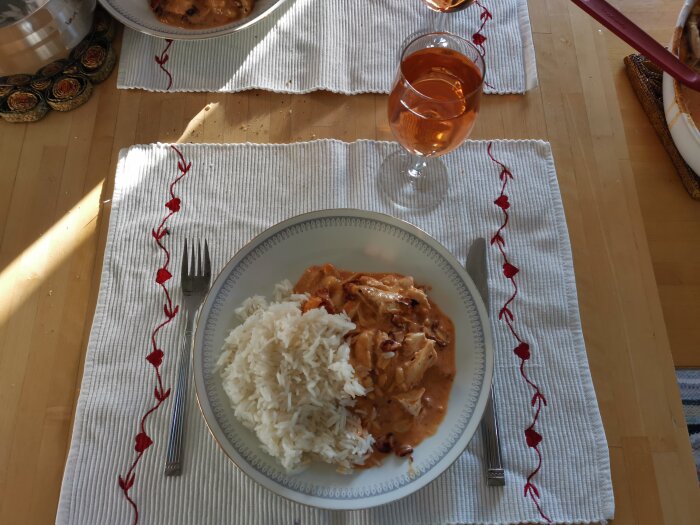 Middagsdukning med vit tallrik, ris, kyckling, gräddsås, glas rosévin, dekorerade bordstabletter, solbelyst rum.