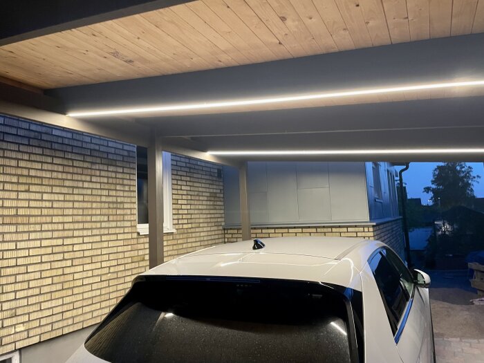 Carport med träpaneltak och LED-belysning, parkerad bil, tegelvägg, skymning.