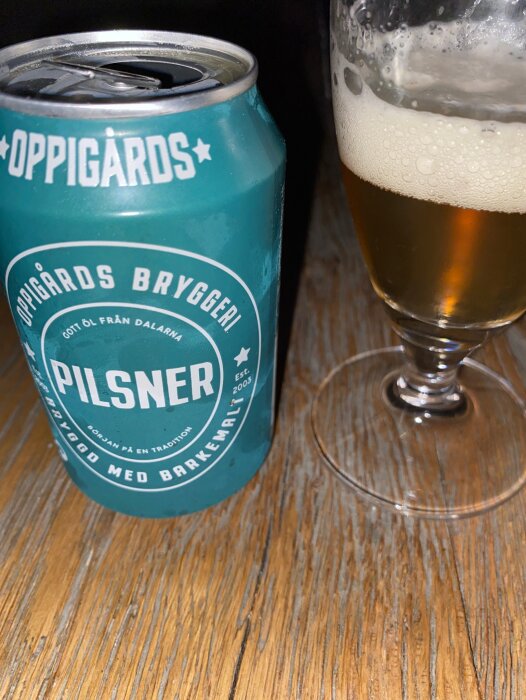 Öppnad burk av Oppigårds bryggeri Pilsner bredvid ölglas på träbord.