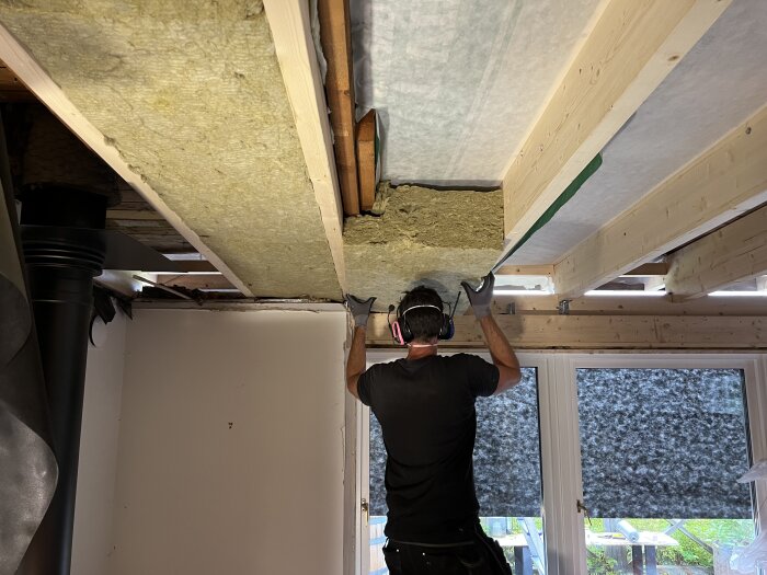 Person installerar isolering i ett takunderlag, bär skyddande hörlurar och handskar, inomhusarbete pågår.