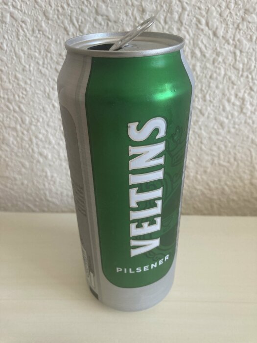 Öppnad aluminiumburk med märket "VELTINS". Grönt och silverfärgat tema. Pilsner. Dryckesförpackning.