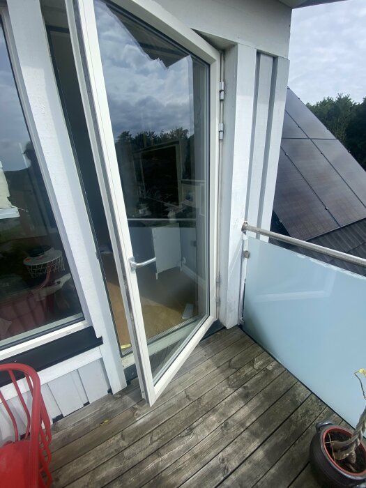 Balkong med öppen dörr, trägolv, glasräcke, reflektion av moln, planterad kruka, urban utsikt.