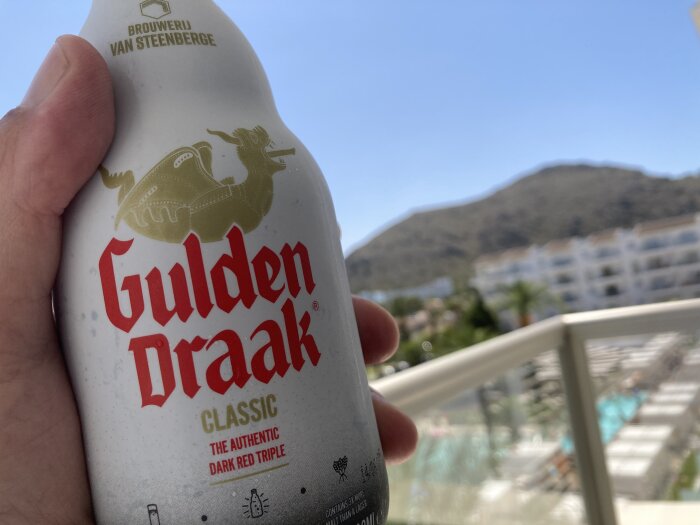 Ölflaska, "Gulden Draak", i fokus, suddig bakgrund med berg, himmel, balkong och byggnader.