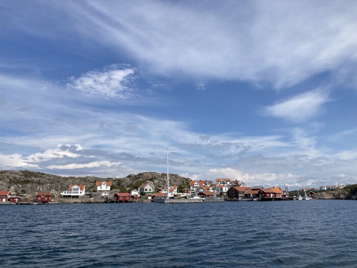 Kustnära svenskt samhälle med röda hus, båtar, klippor och dramatisk himmel.
