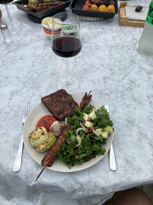 Grillt kött, sallad, grillade grönsaker, vin, utomhusmåltid på marmorbord.